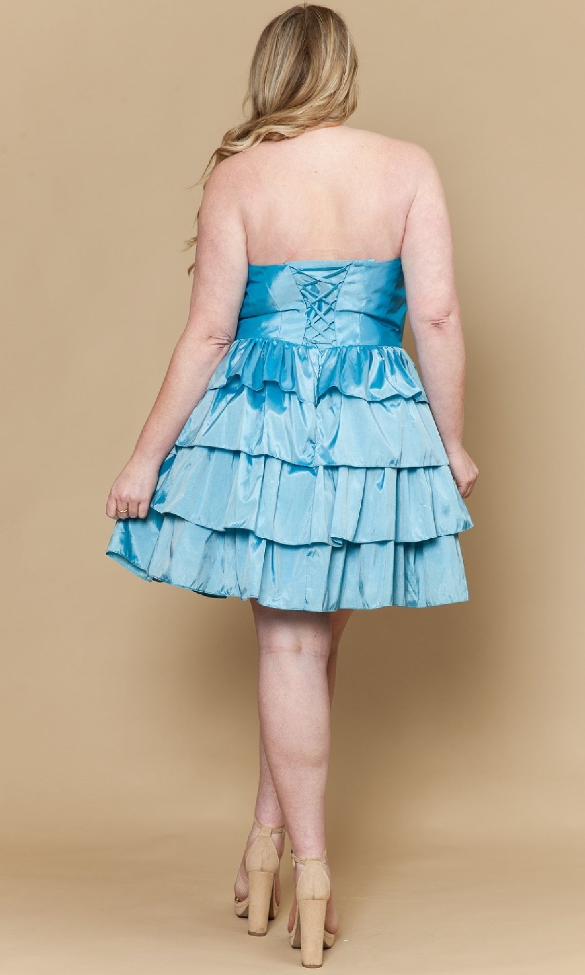 A woman faces away in the Plus-Size Short Halter Party Dress (Poly USA W1216), a light blue ruffled cocktail dress with a lace-up back, paired with beige heels, set against a tan background.
